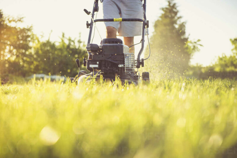 Midsection Of Man Mowing Grassy Field Against Sky In Yard During Sunset